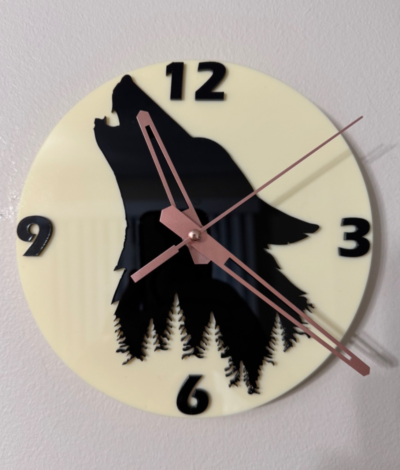 Cream acrylic wall clock with black howling wolf and evergreen silhouettes, black numerals, and rose-toned hands