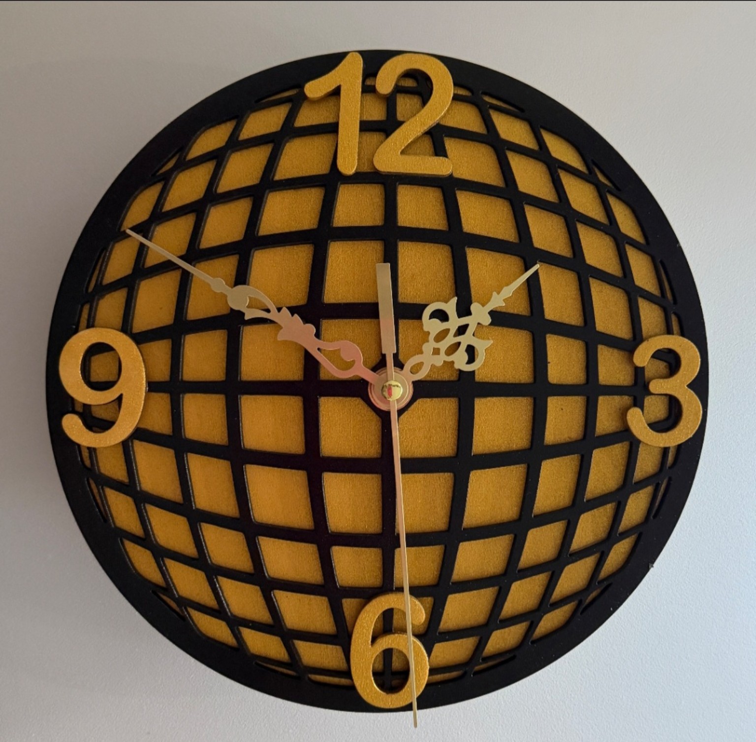 Round black and gold wall clock with curved grid pattern, raised gold numbers and ornate clock hands